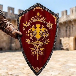 May include: A pointed shield with a deep red center featuring a gold-colored symmetrical floral and heraldic design. The shield has a dark, textured, speckled border that frames the central red panel and the intricate gold-colored ornamentation.