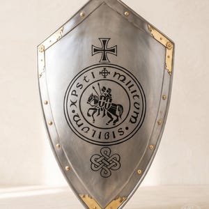 May include: A pointed heater shield made of polished steel with brass corner accents and gold-colored rivets. The center features a black circular emblem depicting a mounted knight, surrounded by the text Sigillum Militum Xpsti.