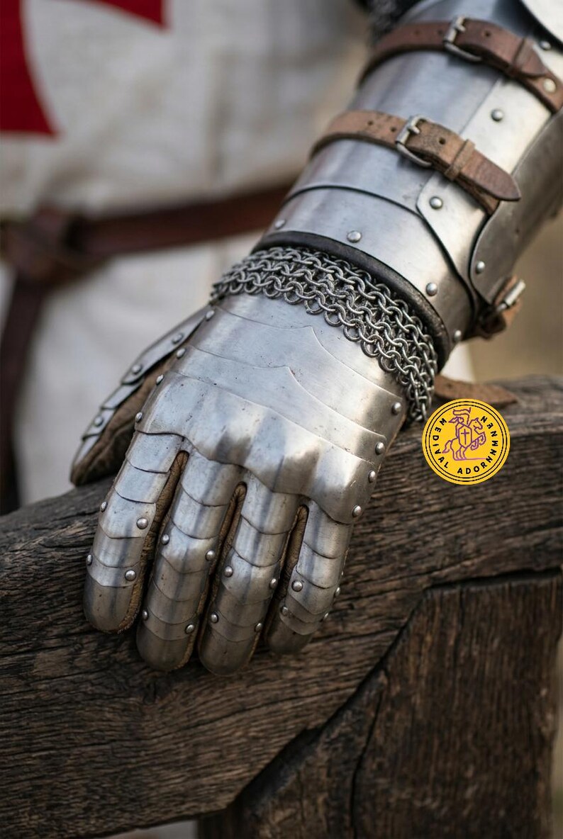 Pu&ograve; includere: Un guanto d'armatura in acciaio con piastre articolate per le dita, un polsino in cotta di maglia e cinturini in pelle marrone con fibbie in metallo. &Egrave; visibile un logo circolare giallo con il testo MEDIEVAL ADORNMENTS e un cavaliere.