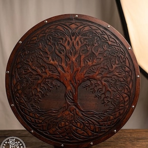 May include: A circular dark brown wooden shield featuring an intricate carved design of a tree with sprawling roots and branches. The outer edge of the shield is accented with evenly spaced silver-coloured metal studs around the entire perimeter.