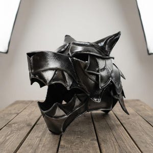 Puede incluir: Un casco con forma de cabeza de lobo negro construido con placas angulares superpuestas. El diseño presenta un hocico puntiagudo, orejas afiladas y una mandíbula abierta con bordes dentados. La superficie tiene un acabado metálico brillante.