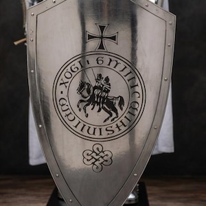 K&ouml;nnte beinhalten: Ein polierter, silberfarbener Metallschild mit schwarzer Gravur. Der Schild zeigt oben ein Kreuz, in der Mitte ein kreisf&ouml;rmiges Emblem mit einem berittenen Ritter und unten ein dekoratives Knotensymbol, umrandet von Nieten am Rand.