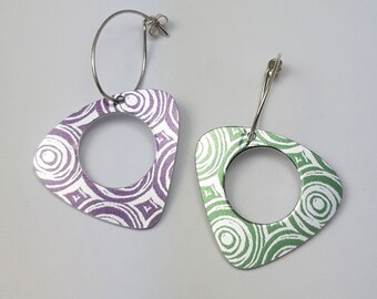 May include: A pair of silver hoop earrings with a geometric design. The earrings are made of metal and have a white background with purple and green swirls.