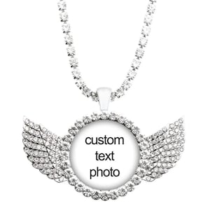 Puede incluir: Un collar con colgante plateado con un centro redondo y detalles de alas, incrustado con cristales transparentes. El círculo central tiene las palabras "custom text photo" en negro. La cadena del collar también está adornada con cristales.