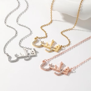May include: Four necklaces with Arabic script pendants. The necklaces are in silver, gold, and rose gold tones. The pendants are encrusted with small, clear crystals. The chains are delicate and the background is white.