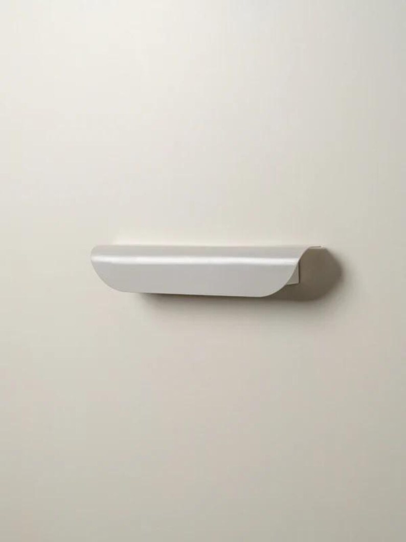May include: A minimalist, light gray floating shelf. The shelf has a curved front edge and is mounted on a neutral-colored wall. The design is simple and modern, suitable for displaying small items or decor.