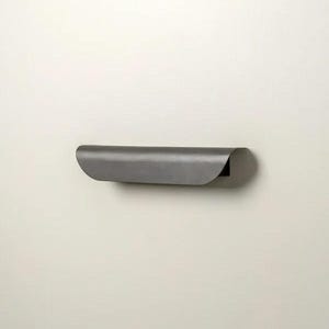 May include: A minimalist, wall-mounted shelf in a dark gray finish. The shelf has a curved, rolled-over front edge and a simple, modern design. It is mounted on a plain, off-white wall.
