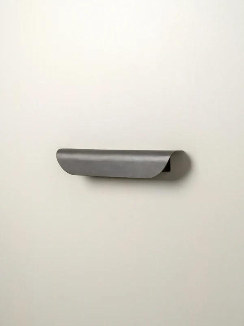 May include: A minimalist, wall-mounted shelf in a dark gray finish. The shelf has a curved, rolled-over front edge and a simple, modern design. It is mounted on a plain, off-white wall.