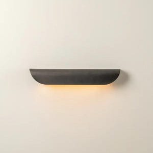 May include: A dark, curved wall sconce with a warm, yellow glow emanating from the bottom. The sconce is mounted on a plain, off-white wall. The fixture has a minimalist design.