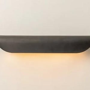 May include: A dark gray, semi-circular wall sconce with a warm, yellow glow emanating from the bottom. The fixture is mounted on a white wall, creating a minimalist design.