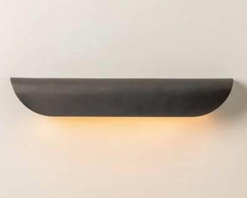 May include: A dark gray, semi-circular wall sconce with a warm, yellow glow emanating from the bottom. The fixture is mounted on a white wall, creating a minimalist design.