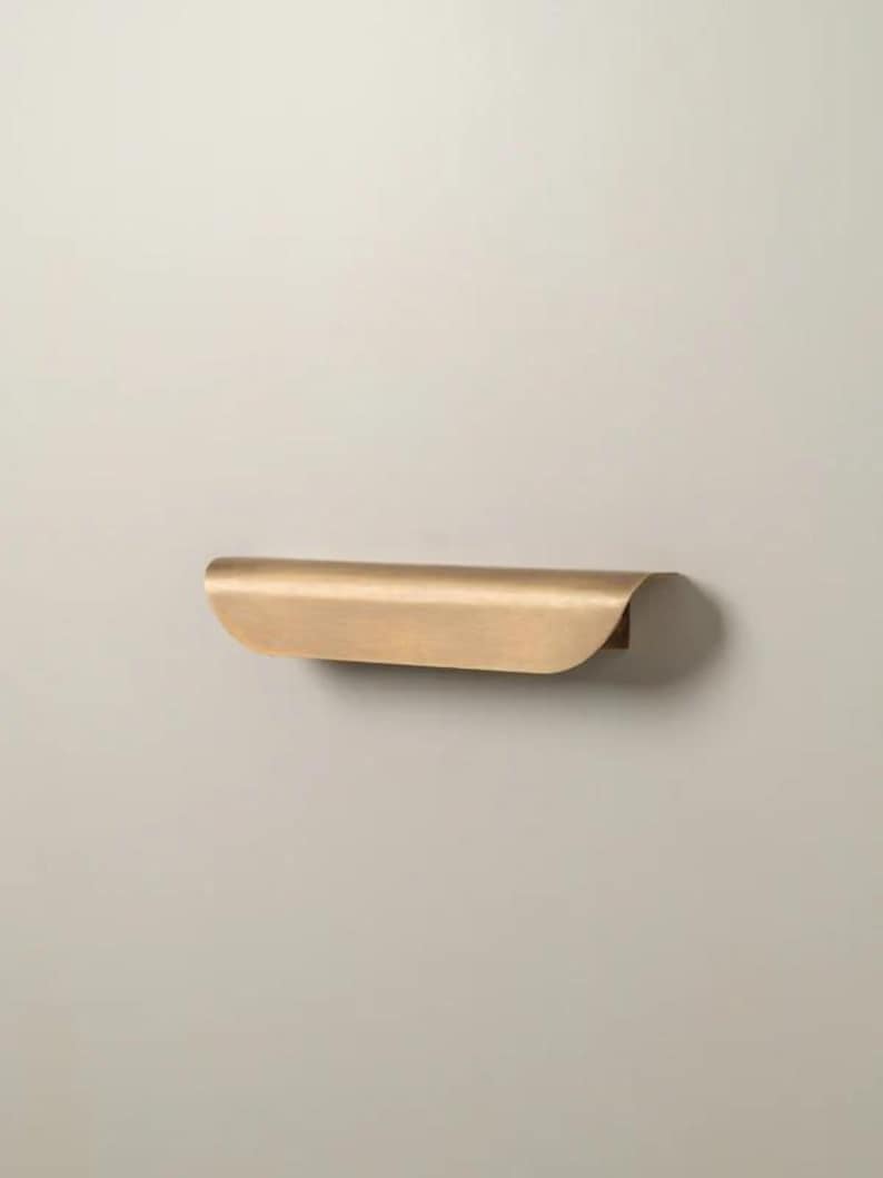 May include: A curved, brass-colored shelf with a rounded front edge and a flat back. The shelf is mounted on a neutral-colored wall. The design is minimalist and modern, suitable for various decor styles.
