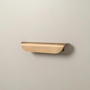 May include: A curved, brass-colored shelf with a rounded front edge and a flat back. The shelf is mounted on a neutral-colored wall. The design is minimalist and modern, suitable for various decor styles.