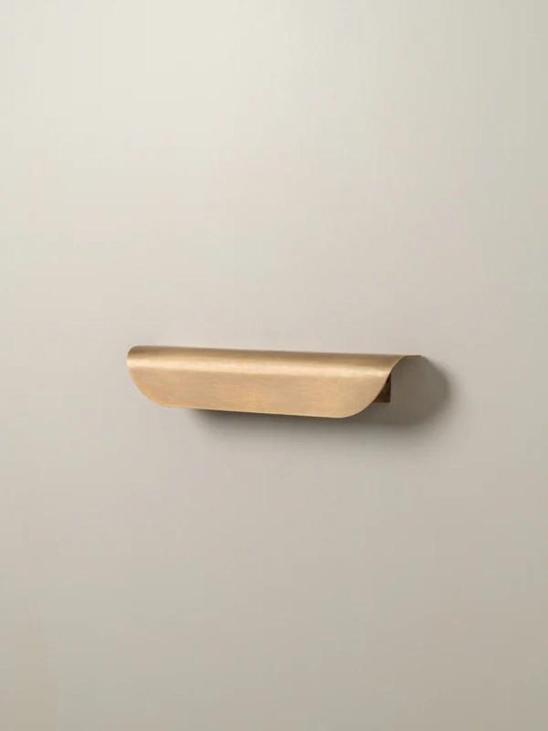 May include: A curved, brass-colored shelf with a rounded front edge and a flat back. The shelf is mounted on a neutral-colored wall. The design is minimalist and modern, suitable for various decor styles.