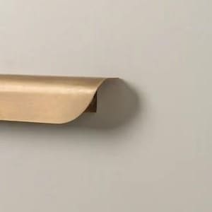 May include: A gold-colored, curved metal shelf with a rounded front edge. The shelf is mounted on a neutral-colored wall. The shelf appears to be made of brass or a similar material.