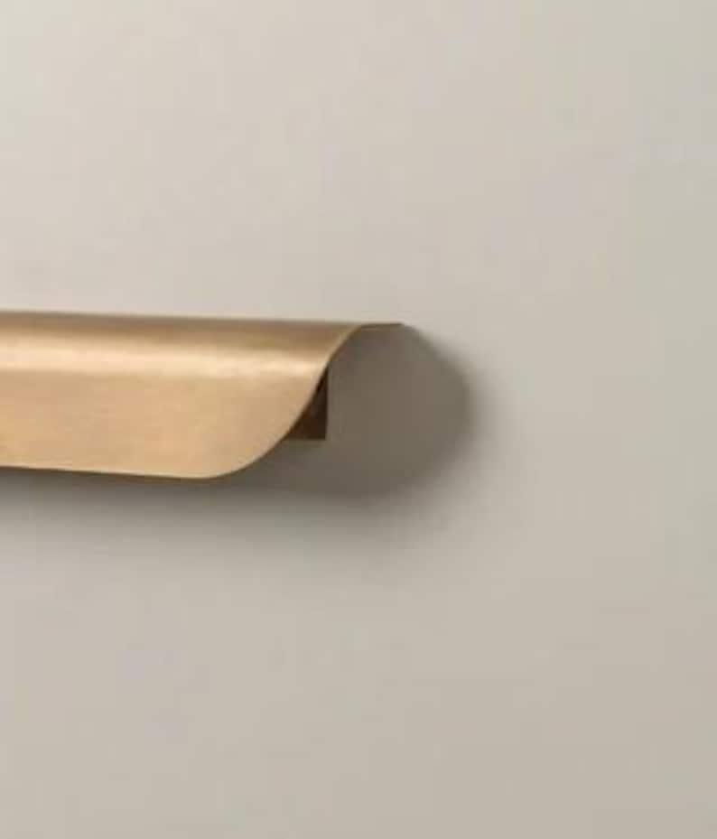 May include: A gold-colored, curved metal shelf with a rounded front edge. The shelf is mounted on a neutral-colored wall. The shelf appears to be made of brass or a similar material.