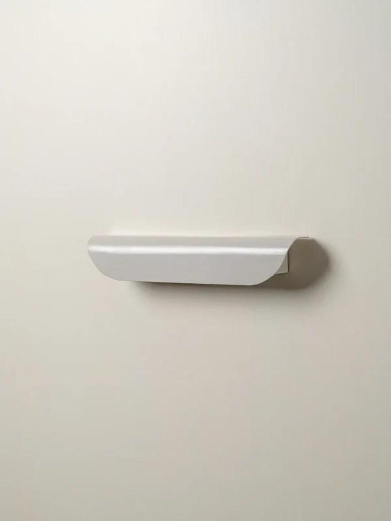 May include: A minimalist, light gray floating shelf with a curved front edge. The shelf is mounted on a neutral-colored wall, creating a clean and modern aesthetic. The shelf is designed for displaying small items or providing storage.