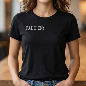 May include: Black crew neck t-shirt with the text "FADE IN:" in white. The t-shirt is worn by a person with brown hair and blue jeans. The person is standing in front of a wooden table.