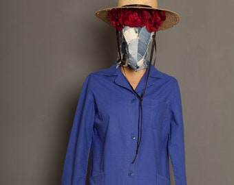 May include: A long-sleeved, blue workwear tunic with a button-up front and two pockets. The tunic is worn on a mannequin with a straw hat and a floral headband.