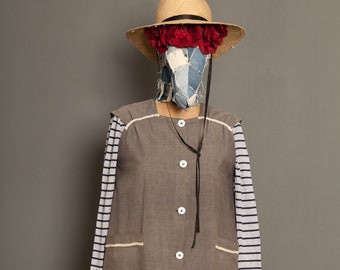 May include: A brown sleeveless dress with white trim and a row of white buttons down the front. The dress has two pockets and is worn over a striped long-sleeved shirt. The model is wearing a straw hat with red flowers.
