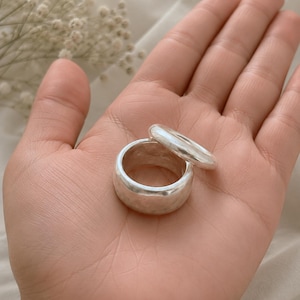 May include: Two silver-toned rings with a smooth, polished finish. One ring features a wide, thick band, while the second ring is thinner and rests partially on top of the wider band. Both rings have a rounded, continuous circular shape.