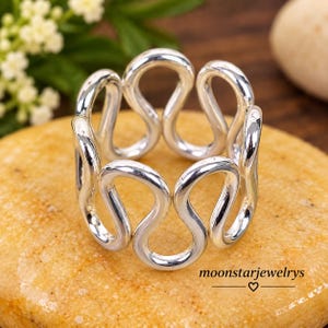 May include: A polished silver ring featuring a continuous, interconnected wavy or serpentine pattern that forms a circular band. The metal surface is smooth and reflective, creating a series of open loops around the circumference of the ring.