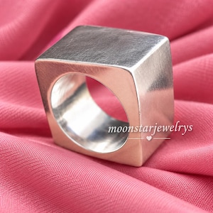 Könnte beinhalten: Ein silberfarbener Ring mit einem markanten quadratischen Außenprofil und einem kreisförmigen Innenband. Die Oberfläche weist eine gebürstete Metallstruktur auf. Der Text moonstarjewelrys ist in Weiß unten rechts auf dem Ring aufgedruckt.