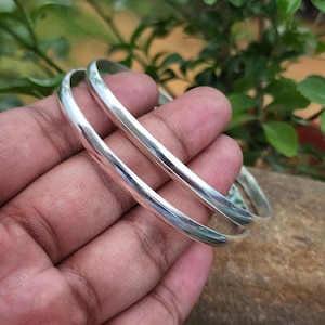 May include: Two shiny, silver bangles are held in a hand. The bangles are smooth and reflective, with a rounded shape. The background is blurred, with green foliage and a brown surface.