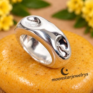 Puede incluir: Un anillo grueso de plata pulida con una superficie lisa y redondeada que presenta dos hendiduras cóncavas. El anillo está colocado sobre una superficie naranja texturizada con el texto moonstarjewelrys y un icono de luna y estrella.