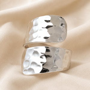 Op de afbeelding: Een zilverkleurige bypass-ring met een brede, open band en een gehamerd, getextureerd oppervlak. Het metaal heeft een reflecterende, gepolijste afwerking die het licht opvangt en duidelijke accenten creëert op het onregelmatige oppervlak.