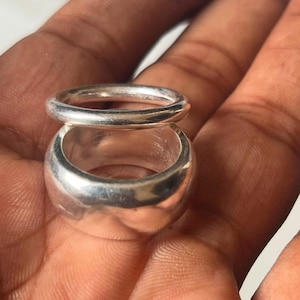 May include: Two polished silver-toned rings stacked vertically. The top ring is a thin, rounded band, while the bottom ring is a wider, thicker band with a smooth, reflective surface. Both rings feature a simple, circular design with no additional embellishments or stones.