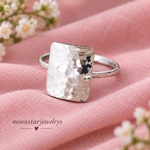 May include: A silver ring featuring a large, rectangular, hammered metal face with a textured, reflective surface. The band is thin and smooth, connecting to the sides of the rectangular centrepiece. The text moonstarjewelrys is visible.