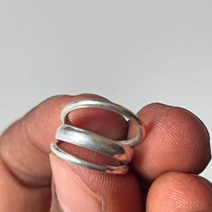 May include: A set of three silver-toned rings with smooth, rounded bands. The rings are stacked together, featuring a polished metallic finish that reflects light. Each band has a simple, uniform circular shape without any additional embellishments.