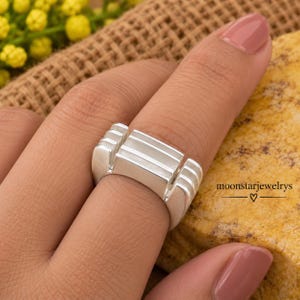 May include: A silver ring featuring a wide, flat-topped band with three distinct horizontal grooves across the centre and vertical ribbed detailing on the sides. The text moonstarjewelrys with a heart symbol is visible on the right side.