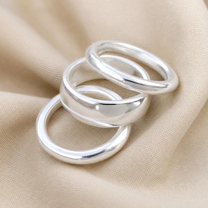 May include: A set of three polished silver rings with smooth, rounded bands. The rings are arranged in a stacked formation, showcasing a reflective metallic finish and a simple, circular design that highlights the clean lines of the jewellery pieces.