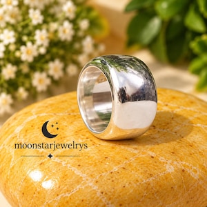Puede incluir: Un anillo de banda ancha de plata pulida con una superficie lisa y redondeada. El anillo está colocado sobre una piedra texturizada de color amarillo anaranjado. El texto moonstarjewelrys está impreso en negro sobre la piedra.