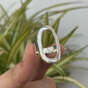 May include: A silver-coloured ring featuring a large, vertically oriented oval-shaped open frame. The metal band is smooth and polished, connecting to the base of the oval structure, creating a bold, open-work design that sits prominently on the finger.