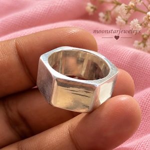 Könnte beinhalten: Ein silberfarbener Ring mit einer markanten hexagonalen Außenform und einer glatten, polierten Oberfläche. Das Band weist flache, kantige Seiten auf, die ein geometrisches Profil bilden und das Licht auf der metallischen Oberfläche reflektieren. Der Text moonstarjewelrys ist sichtbar.