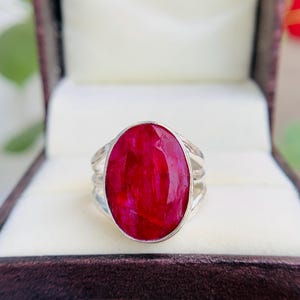 May include: A silver ring featuring a large, oval-cut ruby gemstone. The ruby is a deep, vibrant red color and is set in a simple, elegant silver band. The ring is displayed in a dark brown jewelry box.