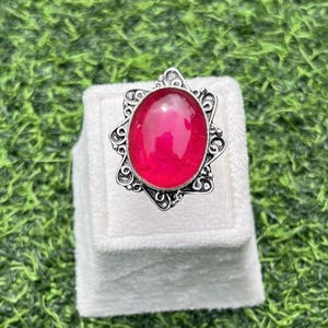 Puede incluir: Un anillo de plata con una gema ovalada de color rojo brillante. El anillo tiene un diseño calado y ornamentado alrededor de la piedra, expuesto sobre un soporte blanco sobre un fondo verde.