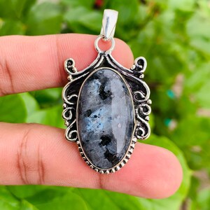 May include: An oval-shaped pendant featuring a dark grey labradorite gemstone with blue iridescent flashes and black inclusions, set in a decorative silver-toned metal frame with intricate scrollwork and a beaded border detail.