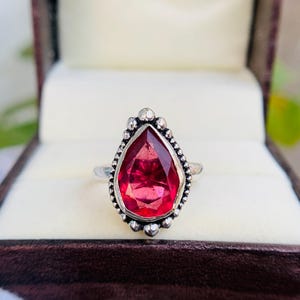 May include: A silver ring featuring a large, teardrop-shaped, faceted red gemstone. The gemstone is surrounded by a beaded silver border. The ring is presented in a brown jewelry box with a cream-colored interior.