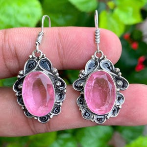 Op de afbeelding: Een paar hangende oorbellen met ovaal geslepen roze edelstenen in bewerkte zilverkleurige metalen frames met donkere geoxideerde details. Elke oorbel hangt aan een eenvoudige zilverkleurige haak en heeft een gefacetteerd oppervlak.