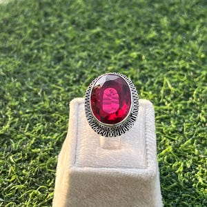 May include: A silver ring featuring a large, oval-cut, ruby-red gemstone. The gemstone is set in a decorative, textured silver band. The ring is displayed on a white jewelry stand against a backdrop of green grass.