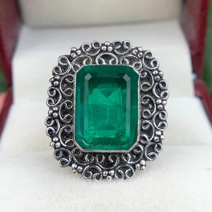 Può includere: Un anello con una grande pietra rettangolare sfaccettata color verde smeraldo incastonata in una montatura in metallo color argento. La montatura è decorata con intricati motivi in filigrana a spirale e piccole perline metalliche.