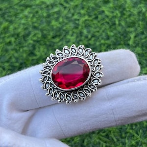 Puede incluir: Un anillo plateado con una gran gema roja rubí de forma ovalada. La gema está engastada en un engaste ornamentado con un diseño floral. El anillo se muestra sobre una superficie blanca sobre un fondo verde.
