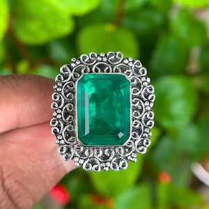 Könnte beinhalten: Ein Silberring mit einem großen, rechteckigen, smaragdgeschliffenen grünen Edelstein. Der Ring hat eine verzierte, filigrane Fassung mit aufwendigen Schnörkeln um den Smaragd. Der Hintergrund ist verschwommenes grünes Laub.