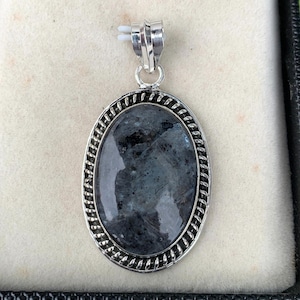 May include: An oval pendant featuring a polished, dark grey stone with mottled inclusions, set in a silver-toned metal frame with a twisted rope border and a simple bail at the top for attachment to a chain or cord necklace.