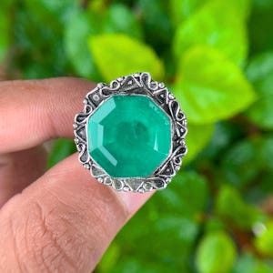 May include: A ring featuring a large, octagonal, vibrant green gemstone set in a decorative, oxidized silver-toned metal frame with intricate scrollwork and beaded detailing along the outer edge, showcasing a polished and textured finish.
