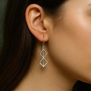 Könnte beinhalten: Silberne geometrische Ohrringe mit zwei verbundenen Diamanten. Die Ohrringe hängen an einem dünnen Haken. Das Design ist modern und minimalistisch. Geeignet für den täglichen Gebrauch.